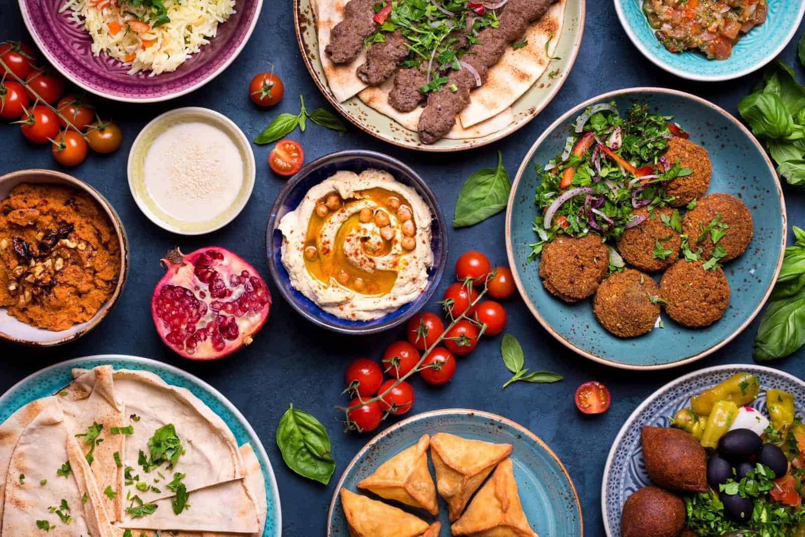 Vista aérea de un festín de comida árabe y de Oriente Medio sobre un fondo oscuro. La mesa está llena de diversos platos coloridos que incluyen falafel crujiente con ensalada, hummus adornado con garbanzos y aceite de oliva, brochetas de carne (kofta) sobre pan plano, samosas doradas, cuencos con salsas, granada abierta, tomates cherry frescos y pan de pita.
